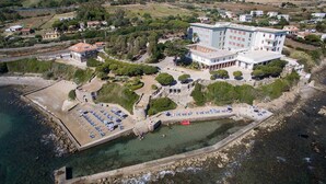 An aerial view of a coastal hotel with multiple buildings, a parking area, and a beach with sun loungers. - Villa MG Hotel (Santa Marinella)