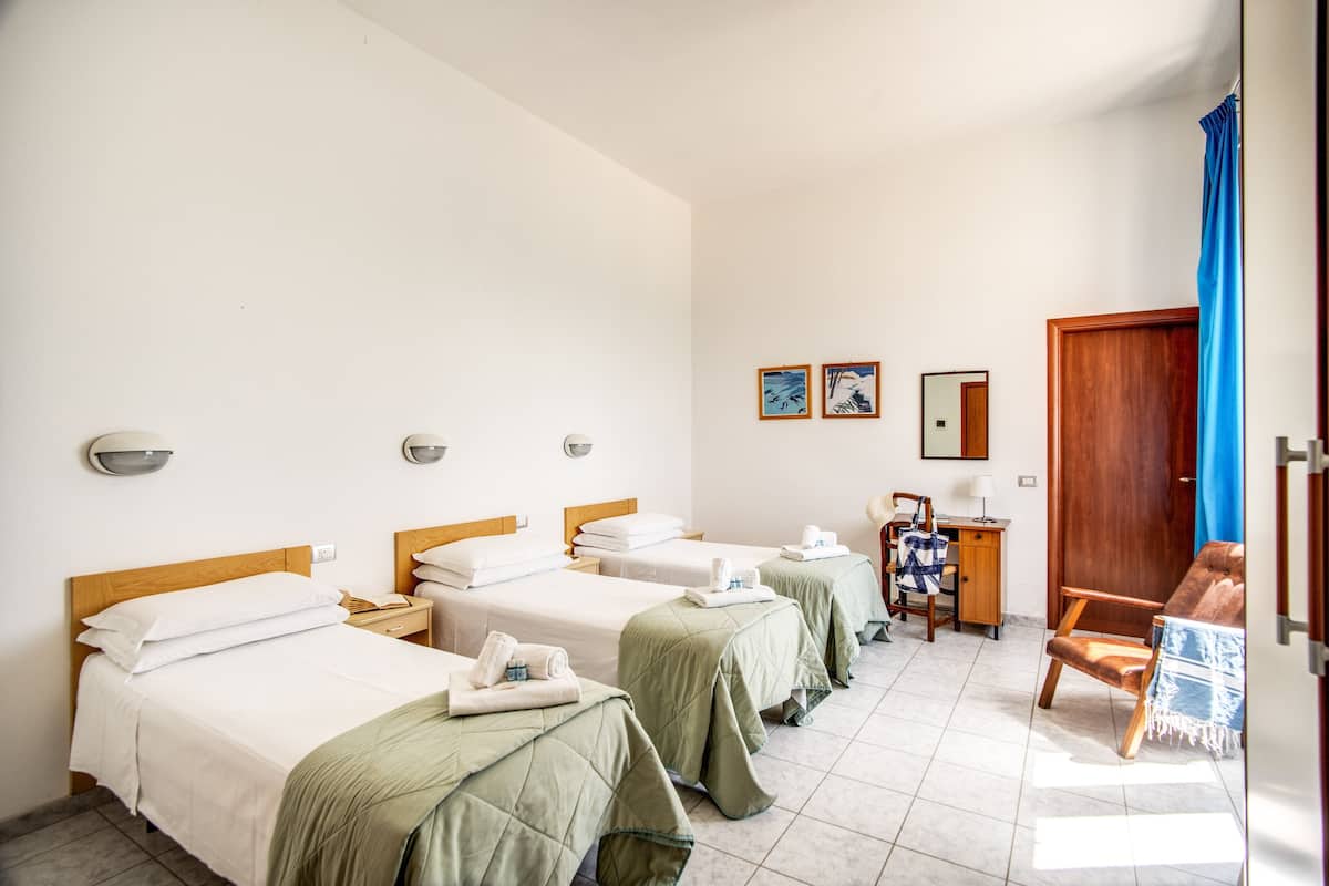 A hotel room with three single beds, a wooden desk, a chair, and a small table with a lamp.
