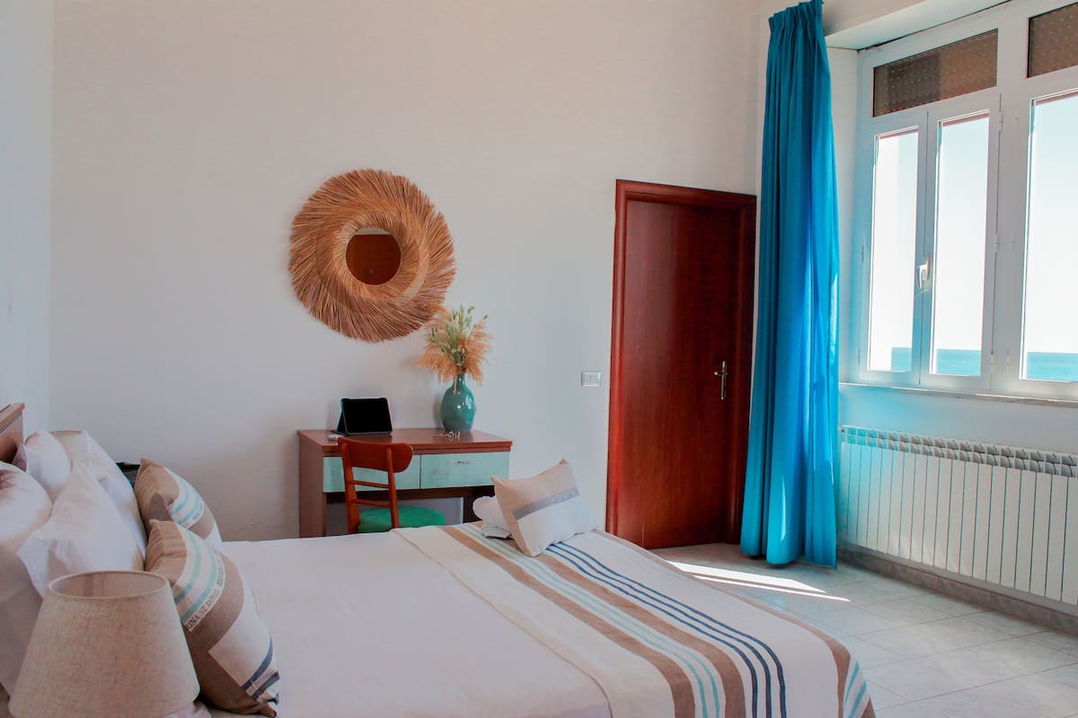A bedroom with a bed, a desk, a chair, a wall-mounted mirror, and a window with curtains.
