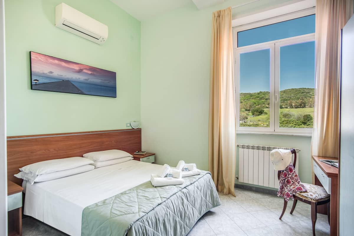 A neatly made bed with white linens, a painting of a landscape on the wall, and a view of greenery through a window.
