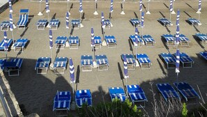 A beach with rows of blue lounge chairs and striped umbrellas, a rocky foreground, and a view of the sea and distant buildings. - Villa MG Hotel (Santa Marinella)