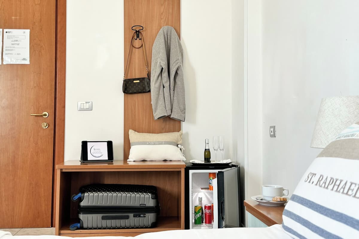 A hotel room with a wooden door, a small desk with a laptop, a suitcase, a minibar, and a bed with a striped pillow.