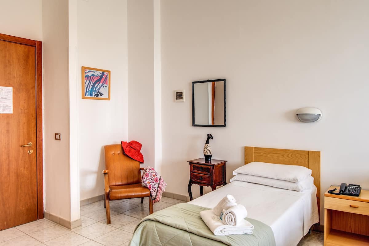 A hotel room with a bed, a chair, a small table, a nightstand, a mirror, and a door.