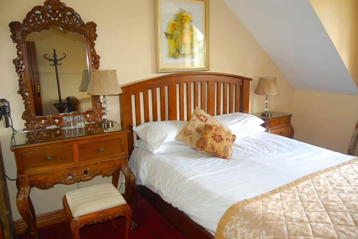 Een slaapkamer met een houten bed, een grote versierde spiegel, een houten dressoir en een ingelijste foto aan de muur.