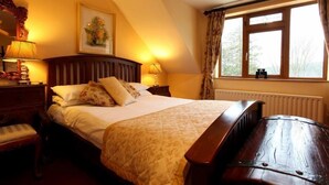 Une chambre à coucher avec un grand lit, une tête de lit en bois, deux tables de chevet avec des lampes, un banc au pied du lit, une fenêtre avec des rideaux et un radiateur.