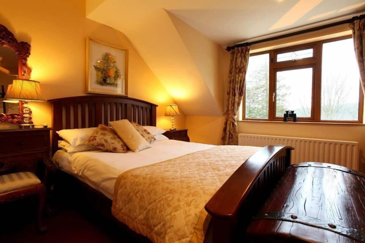 Een slaapkamer met een groot bed, een houten hoofdeinde, twee nachtkastjes met lampen, een bankje aan het voeteneinde van het bed, een raam met gordijnen en een radiator.