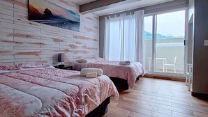 Habitación de hotel con una cama grande, dos camas más pequeñas, un cuadro en la pared y vista al mar a través de una puerta corrediza.