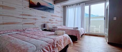 A hotel room with a large bed, two smaller beds, a painting on the wall, and a view of the sea through a sliding door.