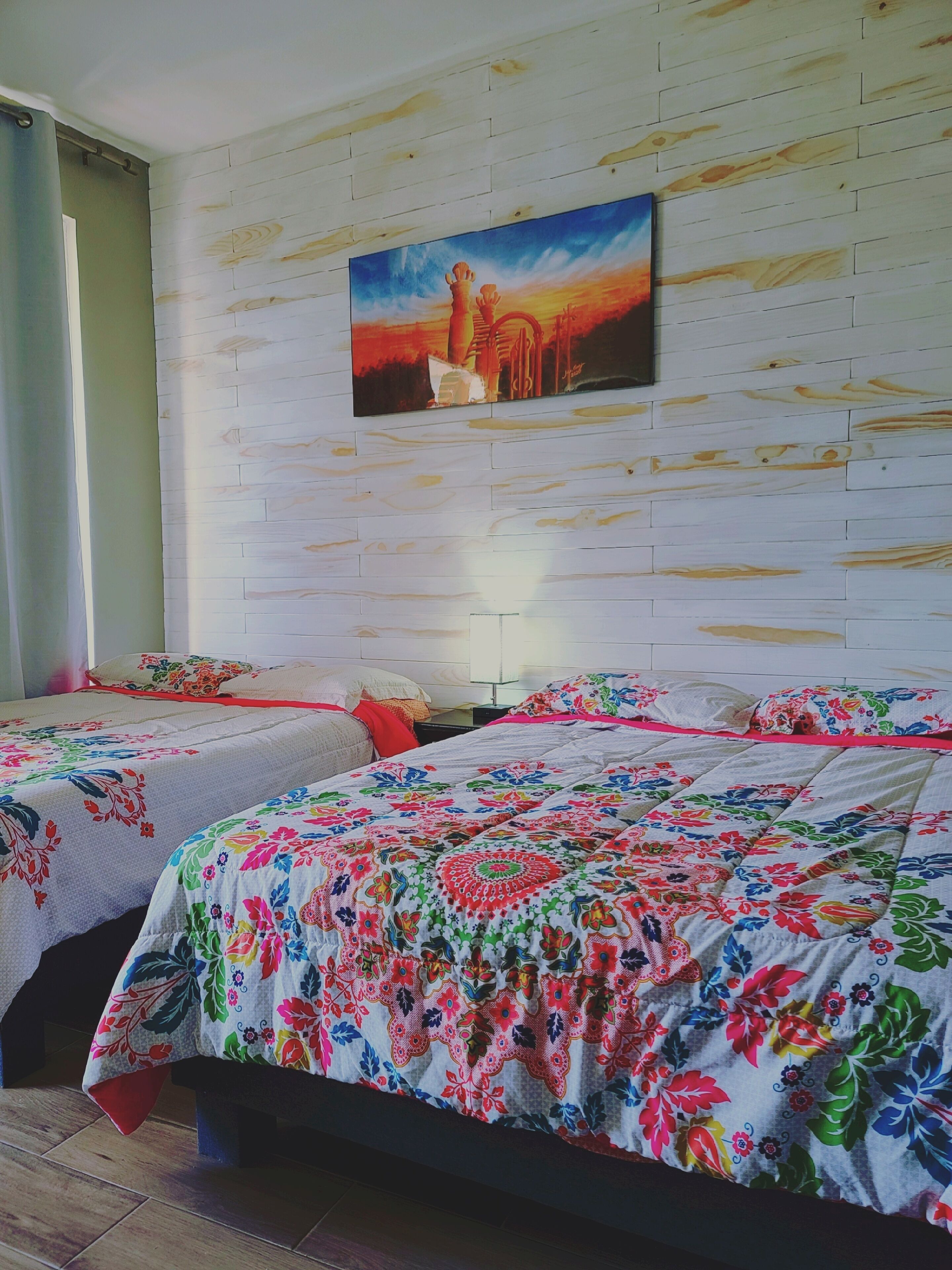 A hotel room with two beds, each with colorful floral bedding. A painting of a sunset is on the wall.