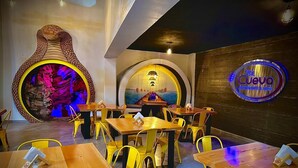 Interior de restaurante con mesas de madera y sillas amarillas, un gran muro decorativo y un letrero de neón.