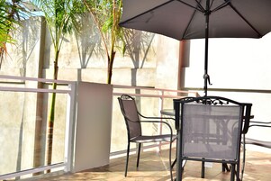 Un balcón con mesa y sillas, un paraguas grande y vistas a palmeras y un edificio.