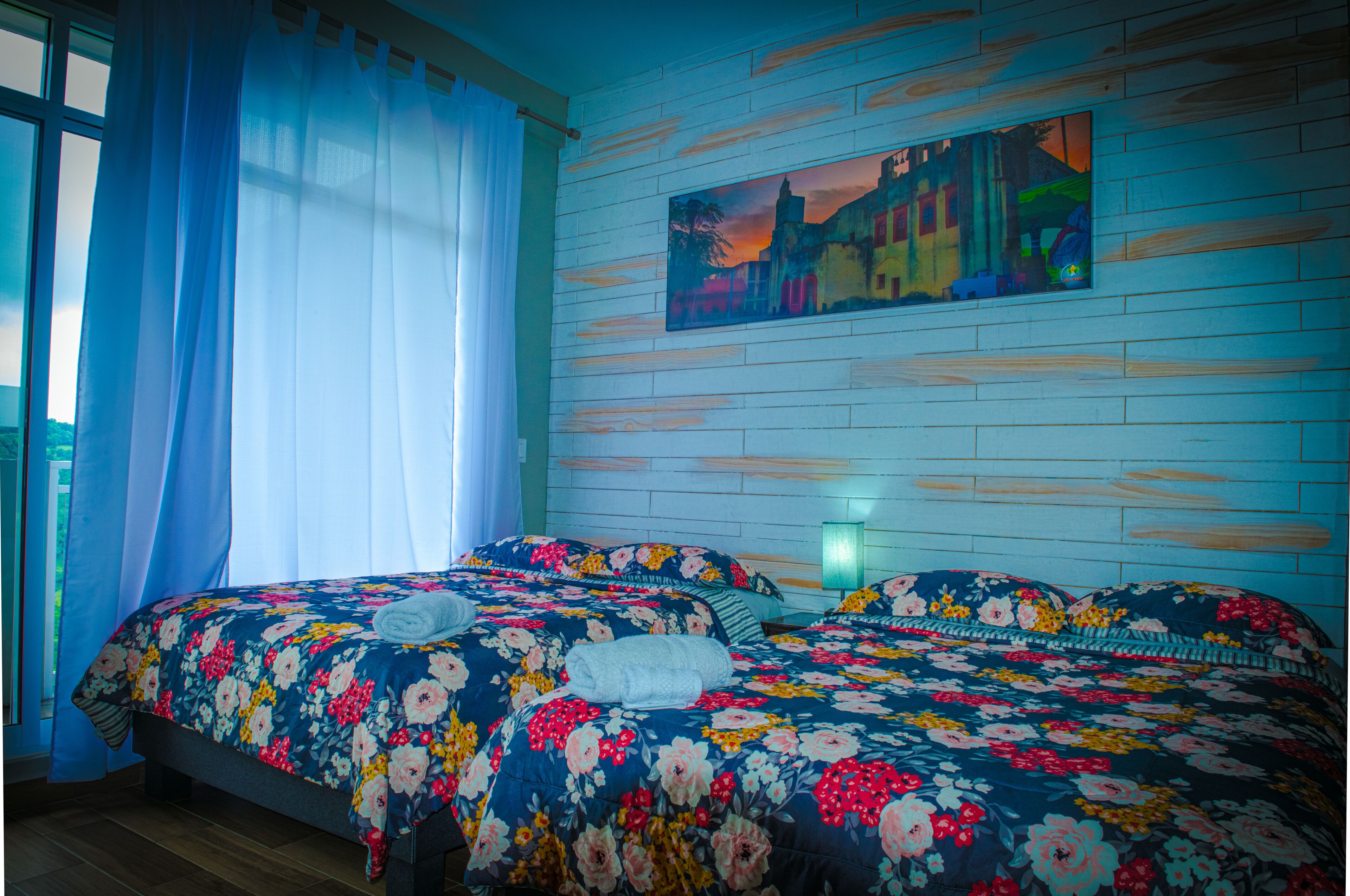 A room with two beds, a window with curtains, a painting on the wall, and a nightstand with a lamp.
