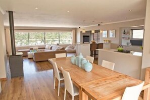 Ein modernes Wohnzimmer mit einem großen Esstisch aus Holz, weißen Stühlen und einer Couch. Es gibt eine Küchenzeile mit Herd und einem Essplatz mit dekorativen Vasen.