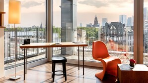 Une chambre moderne équipée d’un bureau, d’une chaise et offrant une vue sur le paysage urbain grâce à de grandes fenêtres.