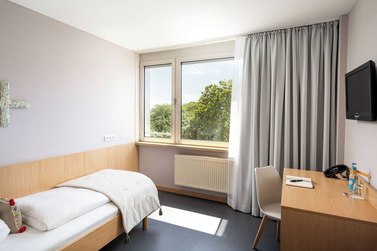 Habitación de hotel con cama, escritorio, silla, televisión, ventana con cortinas y radiador.