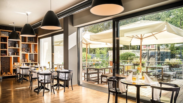 Un espace de restauration intérieur doté d’un plancher en bois, de chaises noires, de tables rondes et de grandes fenêtres donnant sur la terrasse extérieure.
