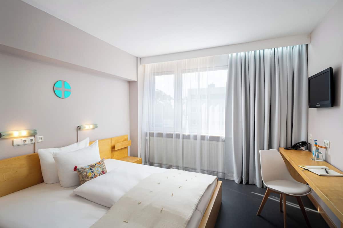 Habitación de hotel con cama, escritorio, silla, televisor y ventana con cortinas.