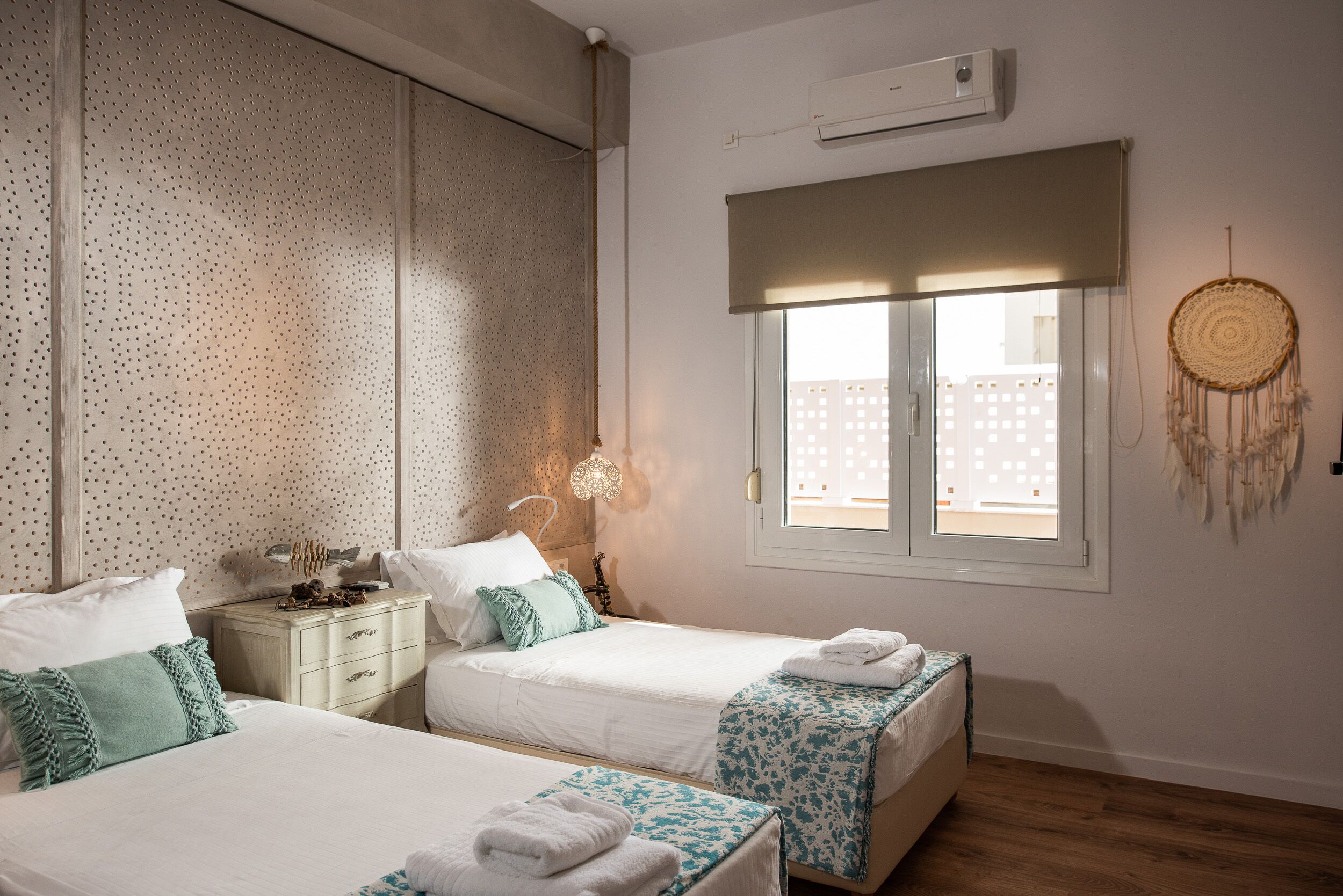 Une chambre avec deux lits, une fenêtre donnant sur un beau paysage, un climatiseur fixé au mur et un attrape-rêves décoratif.