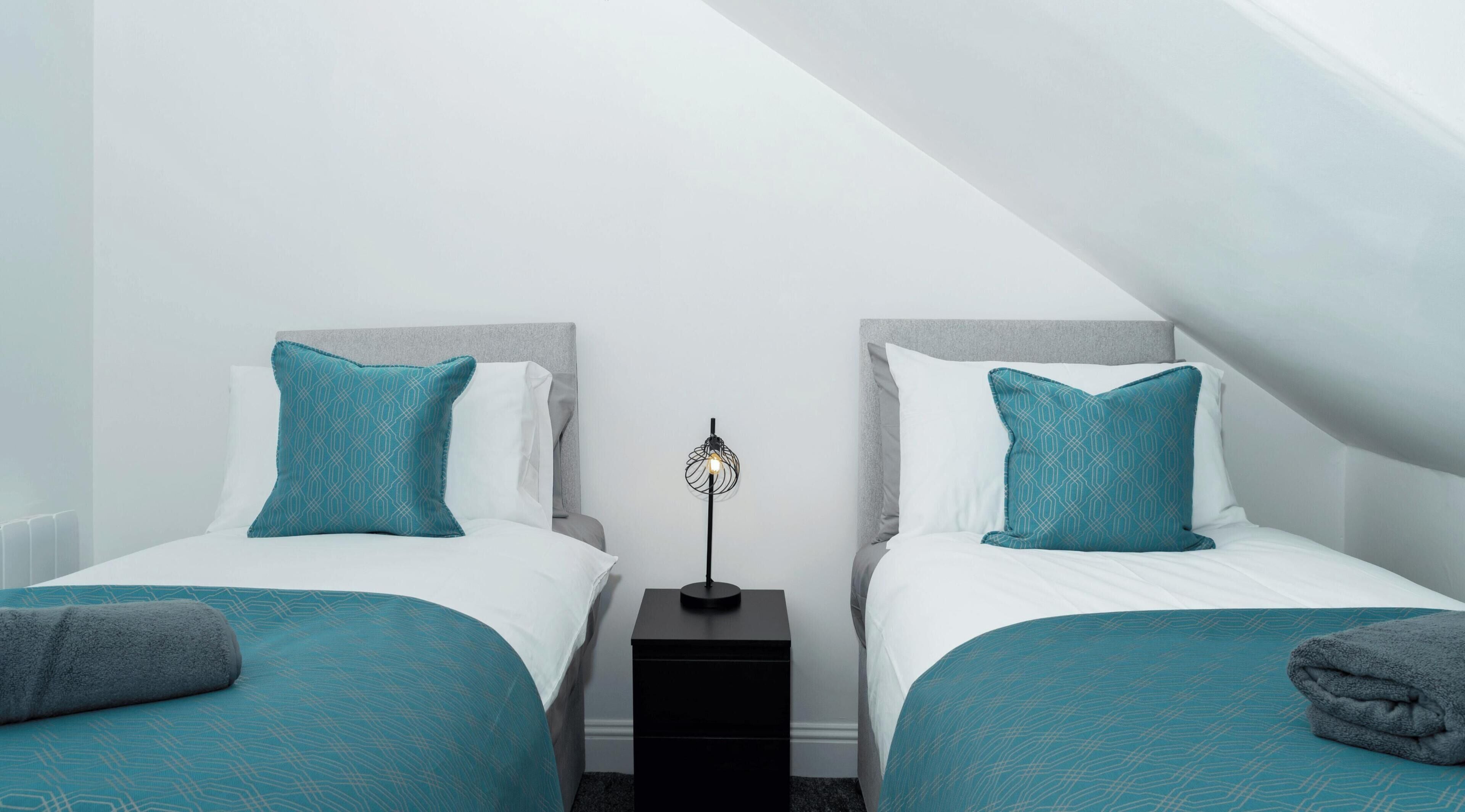 A twin room with two beds, each with teal bedding and matching pillows, a black nightstand with a floor lamp, and a white ceiling.