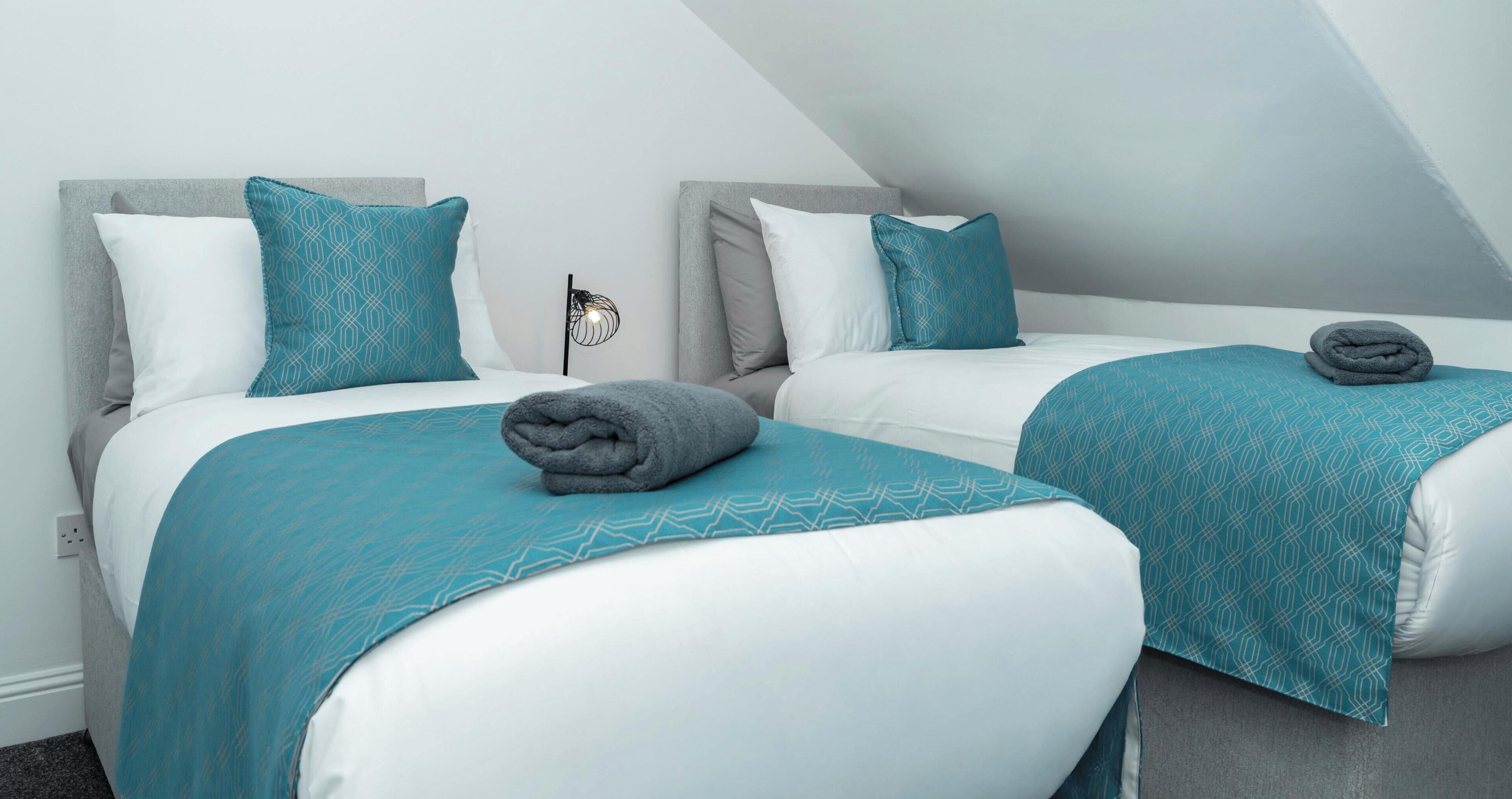 Two twin beds with matching headboards and pillows, a folded towel on each bed, and a wall-mounted light fixture.