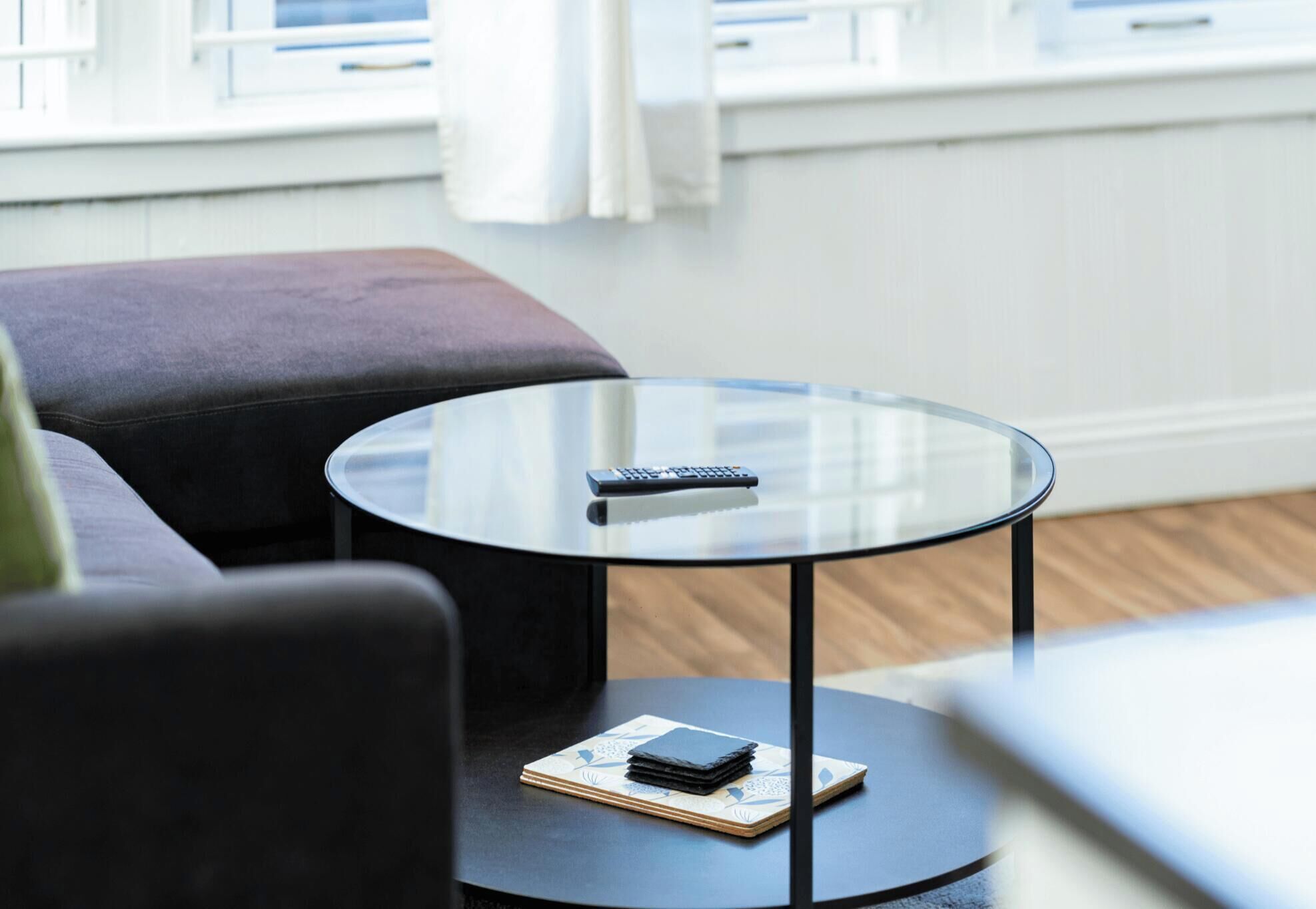 A round glass table with a remote control and a notebook on it, placed on a dark floor near a gray sofa.
