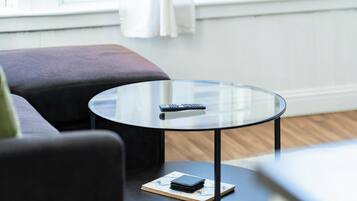 A round glass table with a remote control and a notebook on it, placed on a dark floor near a gray sofa.