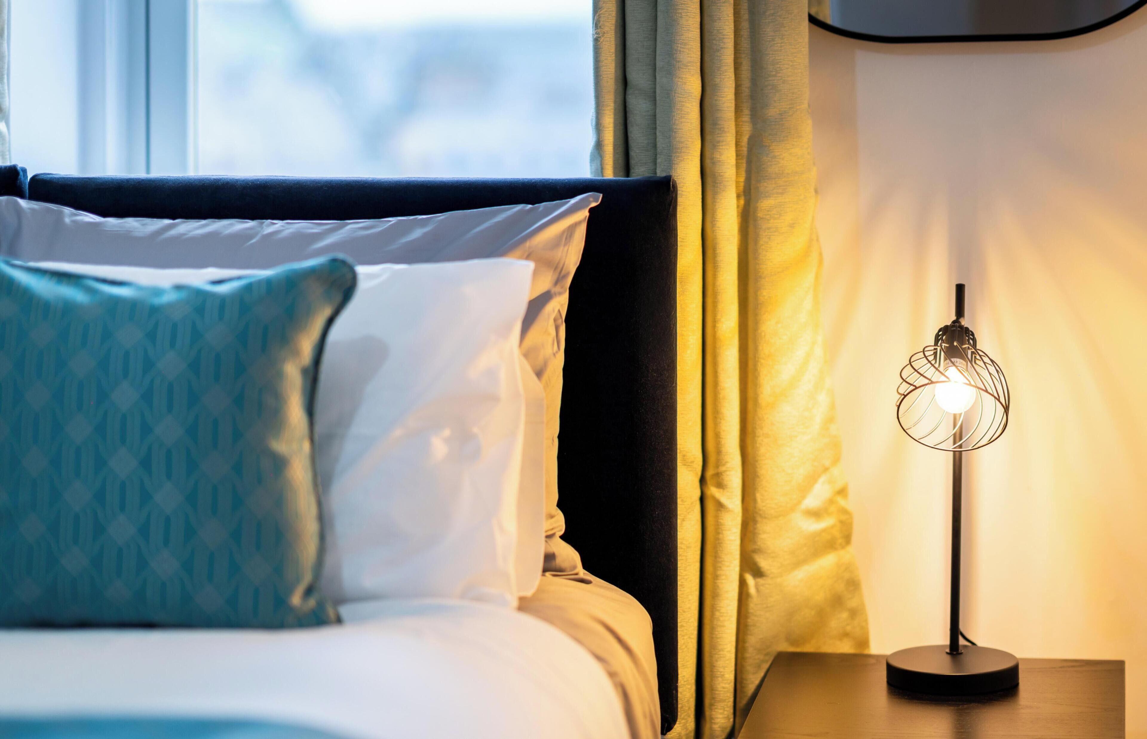 A neatly made bed with patterned pillows, a bedside lamp, and a window with curtains.