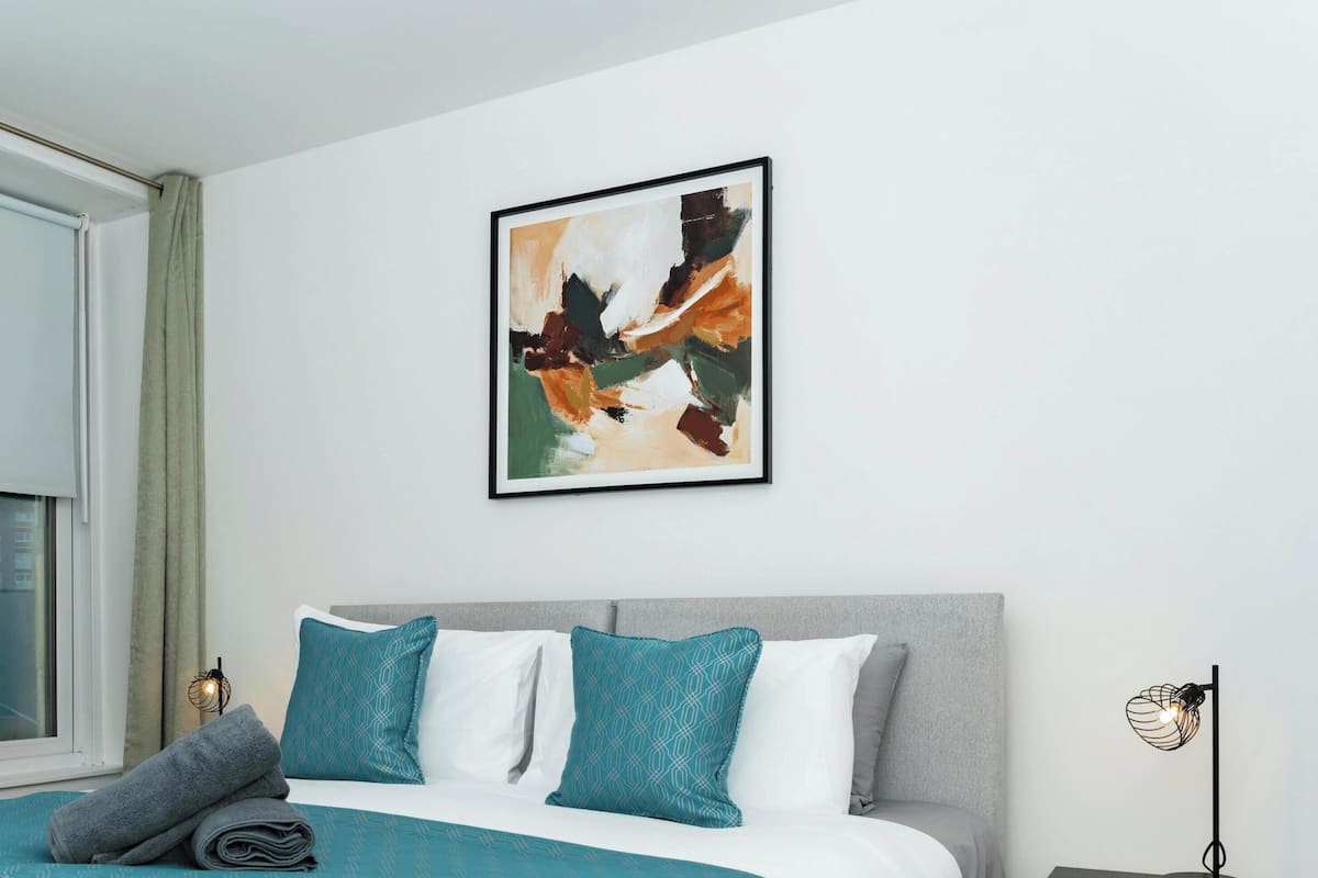 A modern bedroom with a bed, a window with curtains, a wall-mounted abstract painting, and a bedside lamp.
