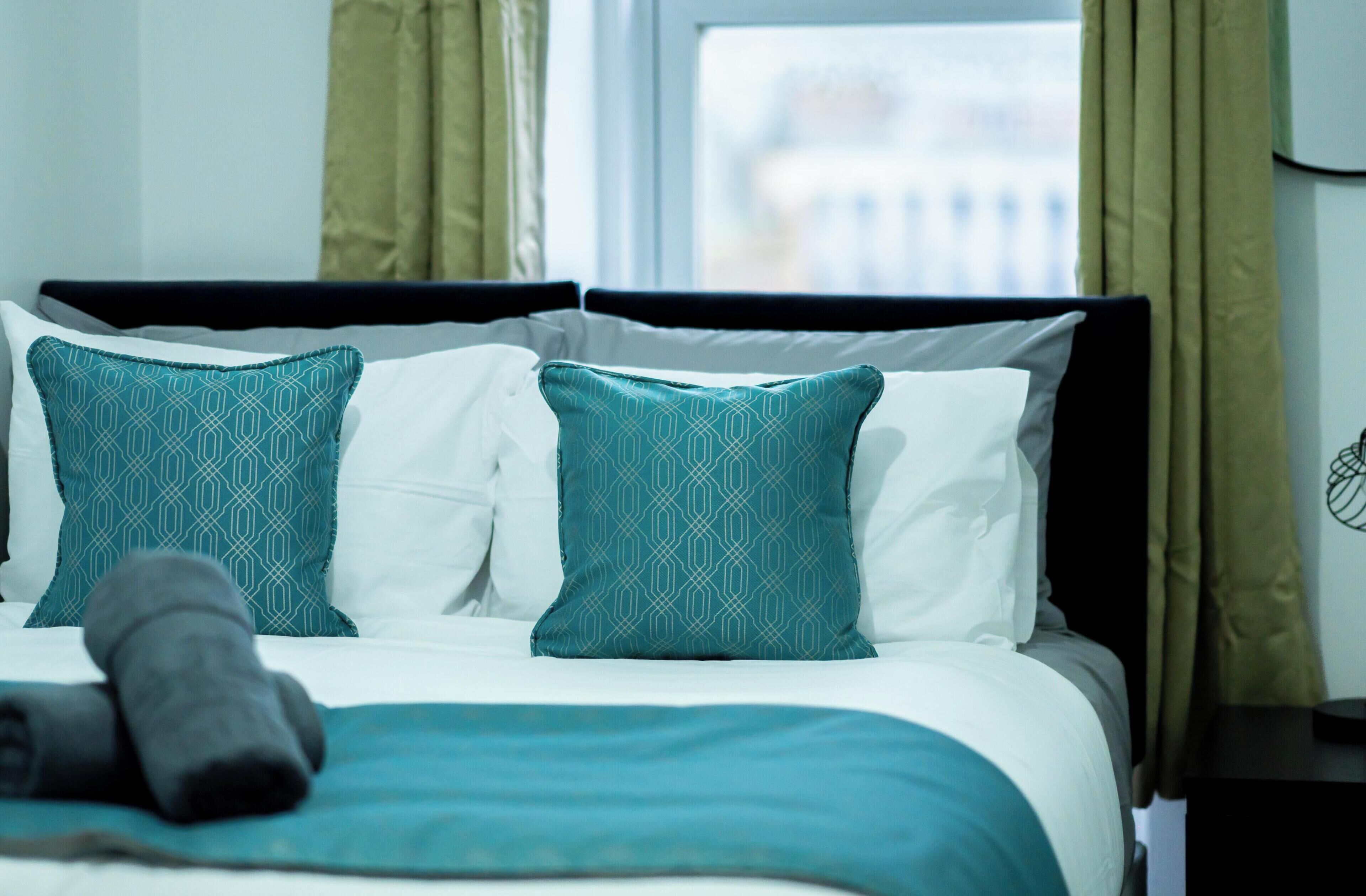 Una cama bien hecha con una almohada de patrón blanco y verde azulado, una manta verde azulado y una toalla enrollada sobre la cama.