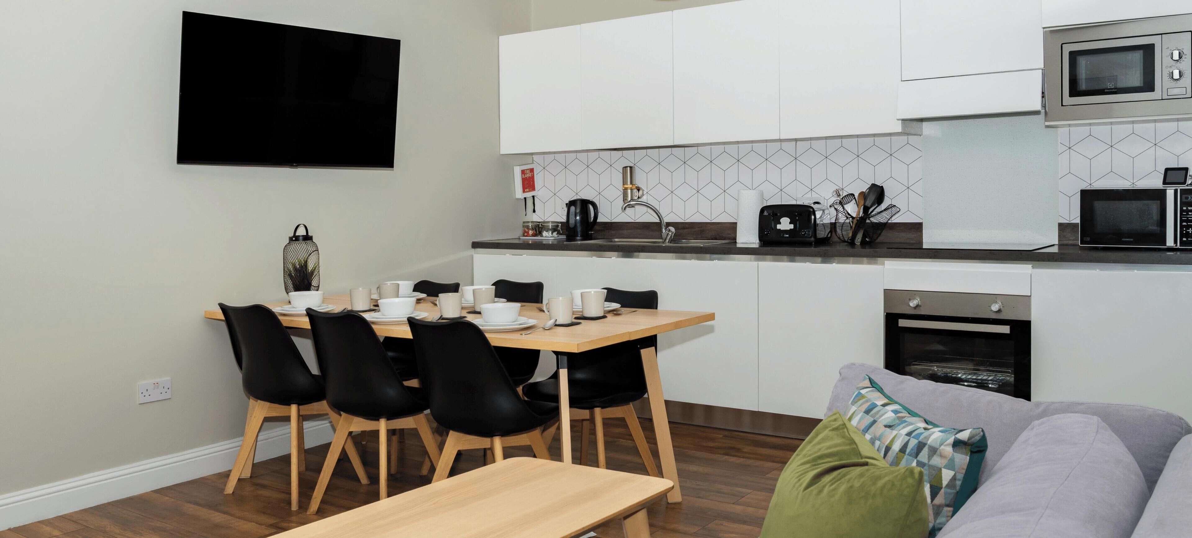 Ett modernt kök med vita skåp, ett matbord med stolar, en platt-TV och en inbyggd ugn.