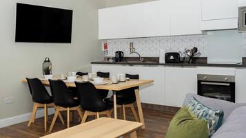 Ett modernt kök med vita skåp, ett matbord med stolar, en platt-TV och en inbyggd ugn.