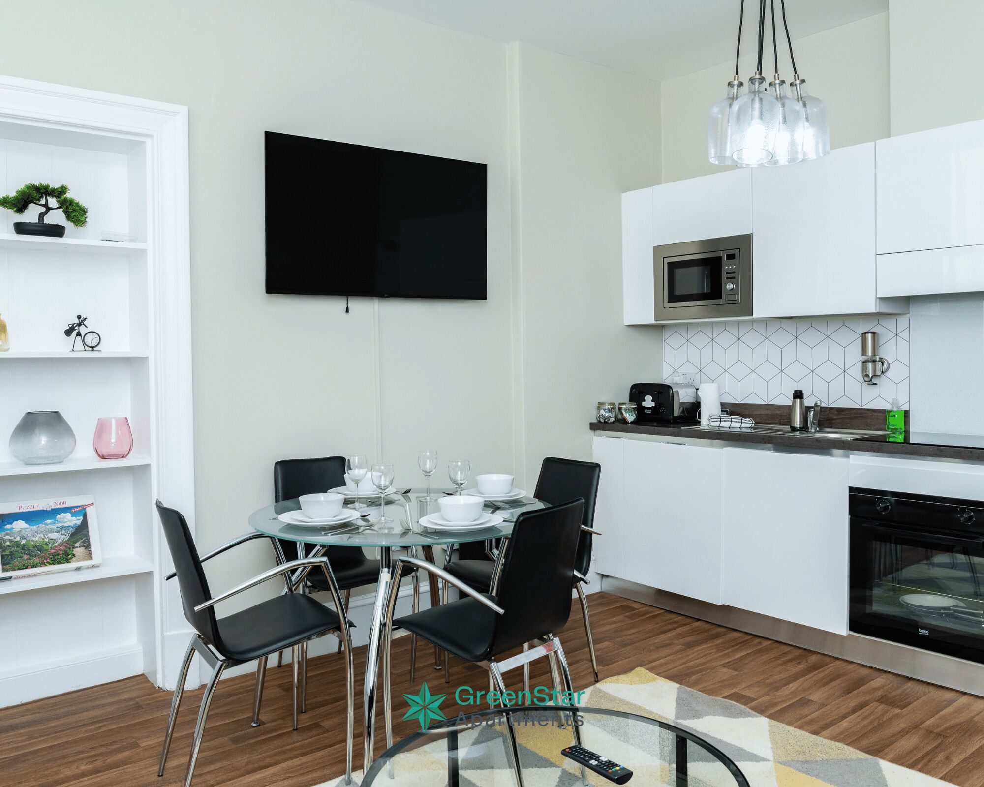 A modern kitchen with white cabinets, a microwave, and a coffee maker. There is a dining table with chairs and a TV mounted on the wall.