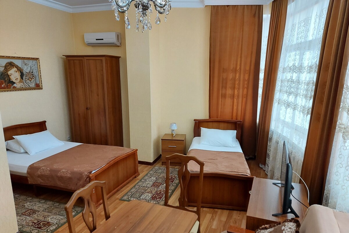 Habitación de hotel con dos camas, una mesa de madera, una silla, una mesita de noche, una lámpara y un televisor.