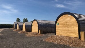 A row of cylindrical wooden structures with a sloped roof, possibly a series of storage or utility buildings. - Cheshire Glamping Pods (Frodsham)