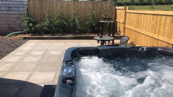 A hot tub with water bubbling, surrounded by a wooden fence, a patio with a table and chairs, and a garden area with plants.