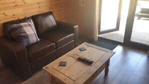 A brown leather sofa with a plaid pillow, a wooden coffee table with two remote controls, and a wooden floor. - Cheshire Glamping Pods (Frodsham)