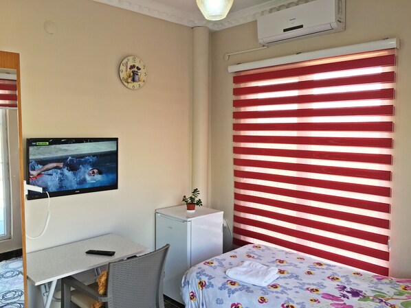 A hotel room with a bed, a small desk with a TV, a refrigerator, a clock, and red-striped blinds. - Dargecit Apart Otel (Dargeçit)