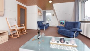 Una sala de estar acogedora con techo de madera, un sillón mecedor, un sofá azul y una mesa de centro de vidrio con copas de vino y velas.