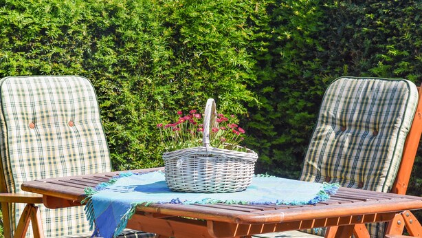Una silla de lounge de madera con un cojín a cuadros, una cesta de mimbre con flores y un paño azul sobre una mesa.