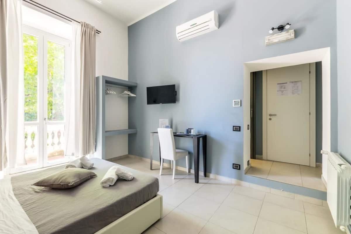 A hotel room with a bed, a desk, a chair, an air conditioner, a TV, and a door.