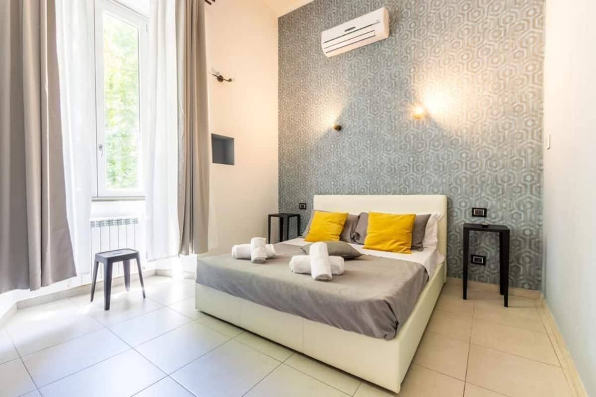 A bedroom with a bed, two bedside tables, a window with curtains, and an air conditioning unit on the wall.