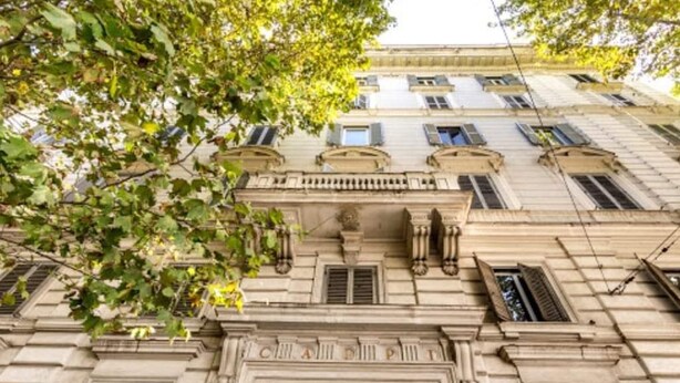 Fachada clásica de edificio con una entrada central, flanqueada por ventanas con contraventanas, y un árbol que enmarca parcialmente la vista.