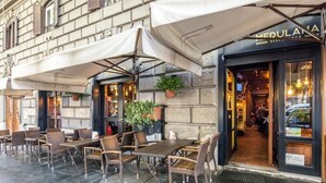 Un café al aire libre con mobiliario de mimbre y sombrillas, fachada de un edificio de piedra y una entrada visible a una tienda.