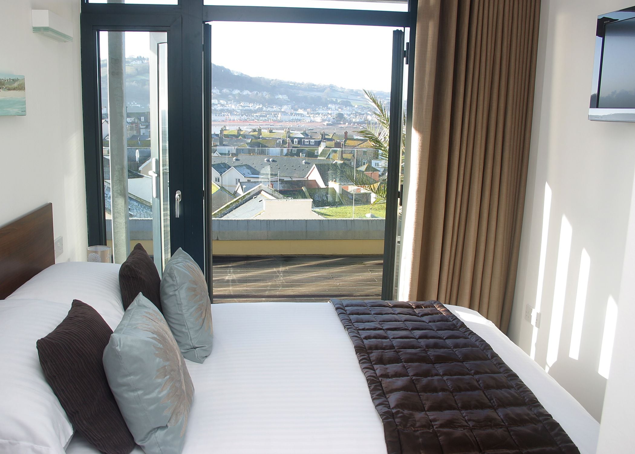 A hotel room with a bed, a view of a town through a glass door, and a television on the wall.