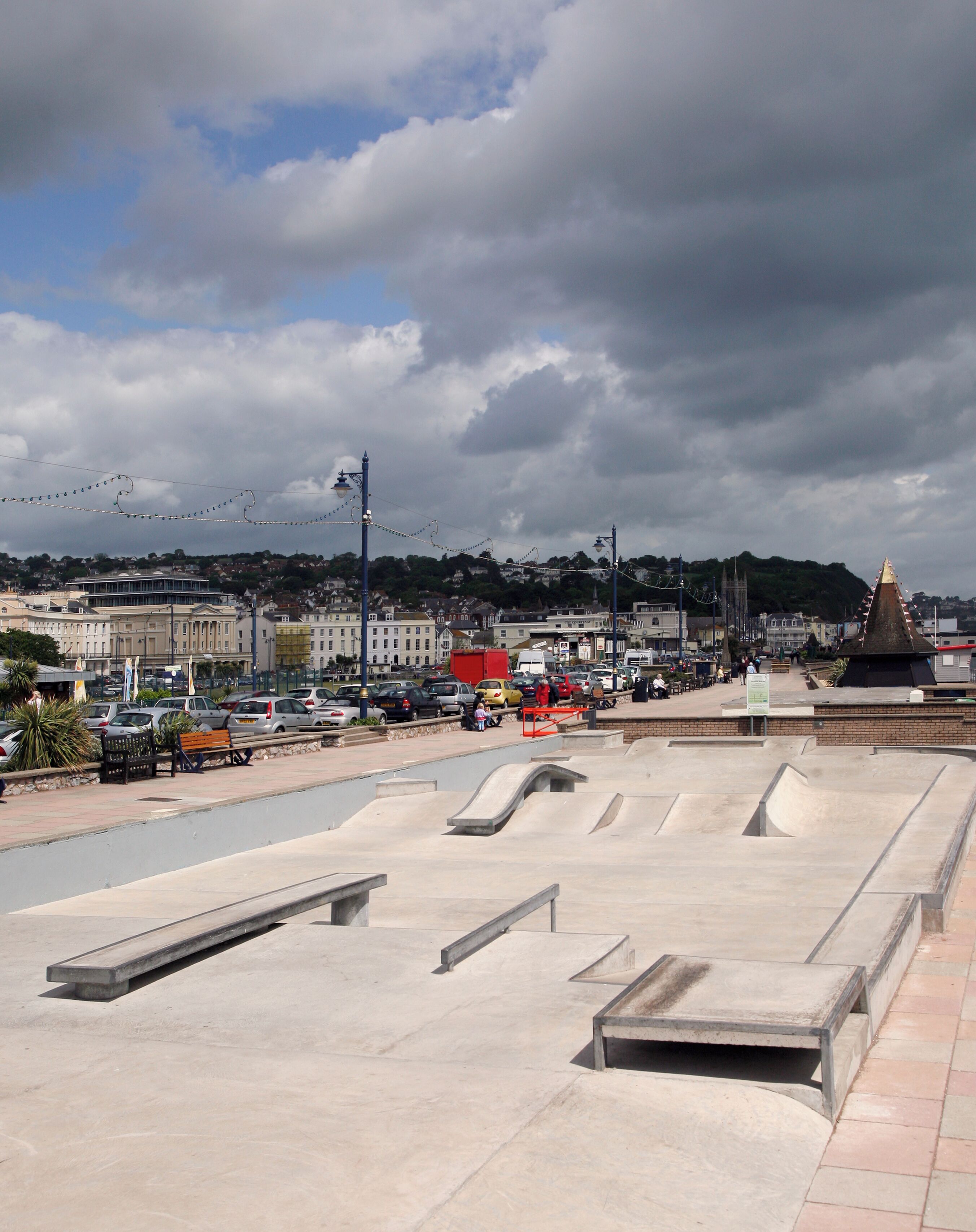 A skate park with ramps and benches, surrounded by buildings and parked cars.