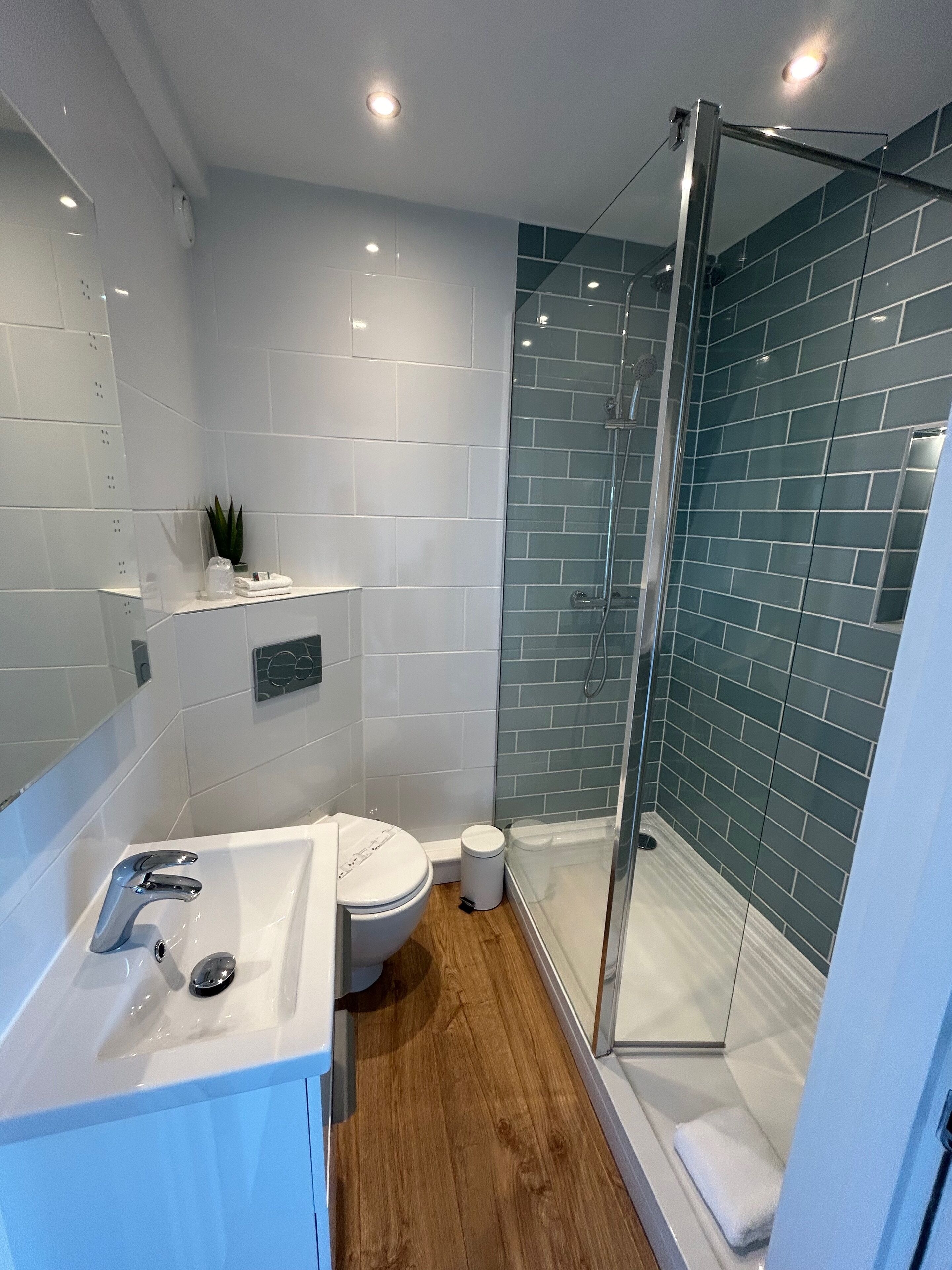 A modern bathroom with a glass-enclosed shower, white tiles, and a wooden floor.