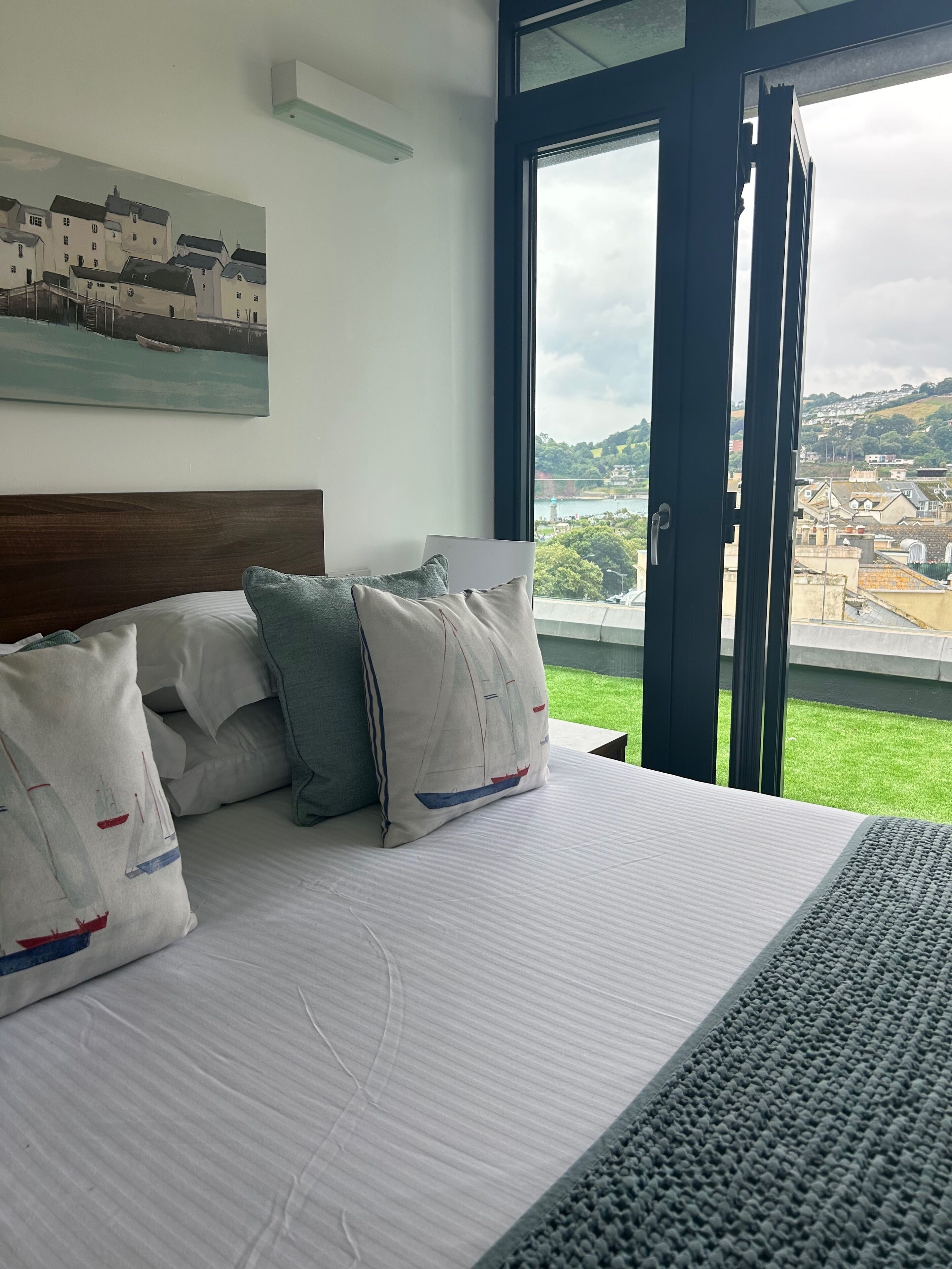 A hotel room with a bed, pillows, a painting, and a large window with a view of a town and greenery.