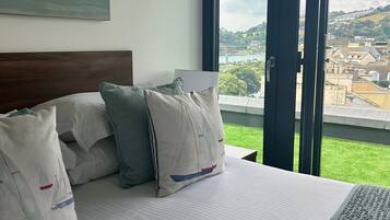 A hotel room with a bed, pillows, a painting, and a large window with a view of a town and greenery.