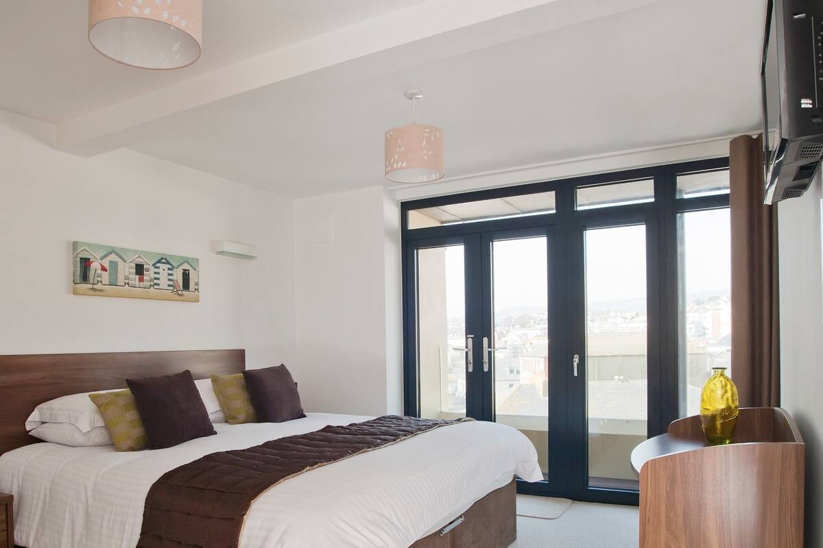 Une chambre avec un grand lit, une table de chevet en bois, un téléviseur et une vue sur l’extérieur, visible à travers une porte vitrée.