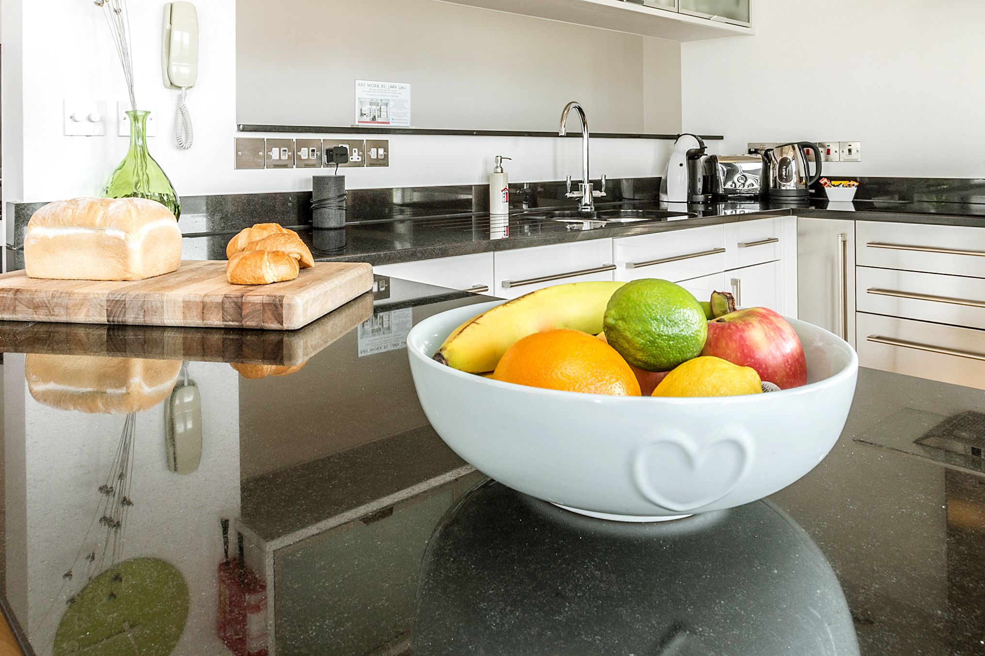 Una cocina con encimera negra, un bol blanco lleno de fruta, una hogaza de pan y una tabla de cortar con una baguette.
